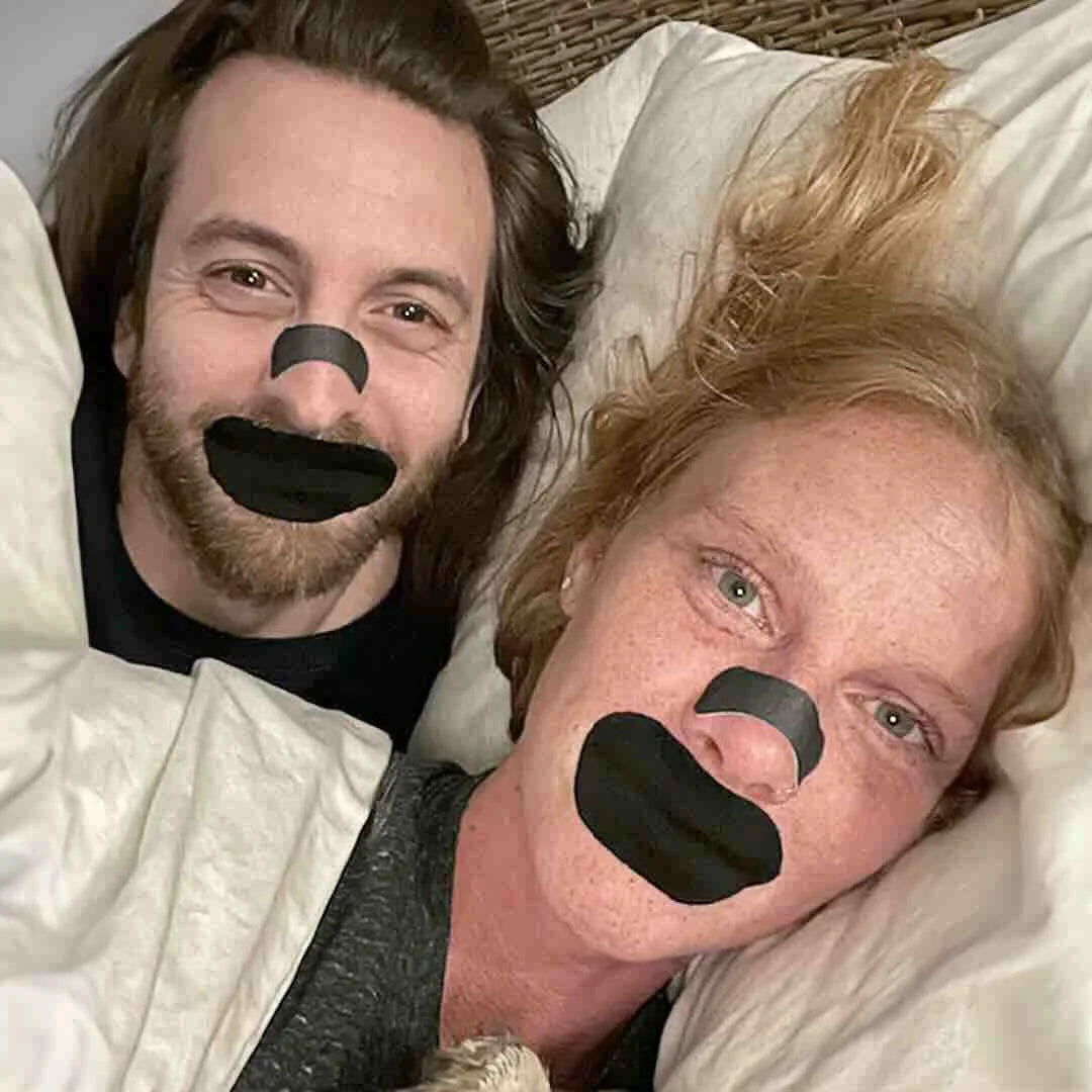 A smiling couple wearing black Hostage Mouth Tape and nasal strips to prevent snoring